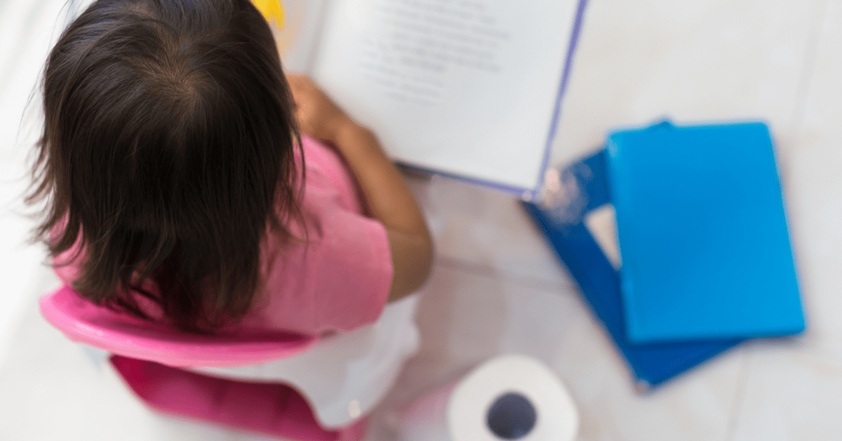 Toddler sitting on a pink potty chair reading a book, with toilet paper and blue books on the floor nearby.