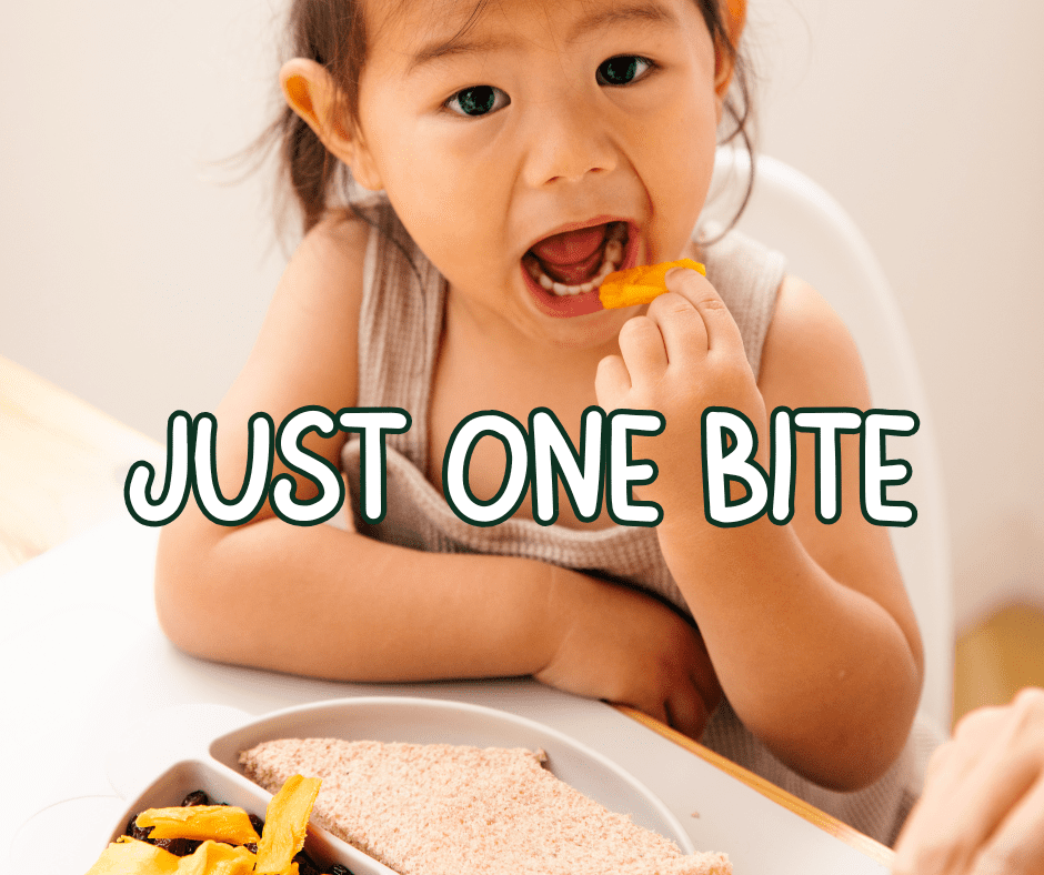 Toddler taking a bite of food at the table, with a plate of berries, dried mango, and a sandwich in front of them. Text overlay reads “Just One Bite.”